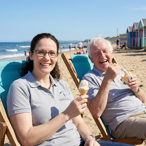 A joyful Silver Skies day out: a companion and elderly client sharing smiles and ice cream on a sunny beach on the glamorgan coast, illustrating flexible support.